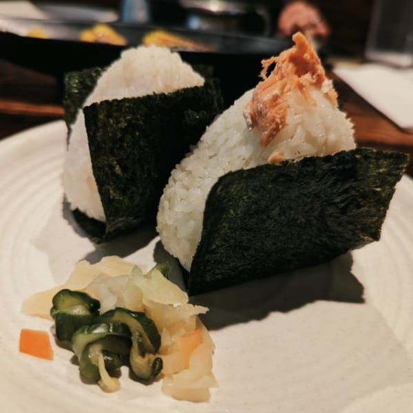 Two onigiri, one topped with salmon, served with pickled ginger and cucumbers on a white plate.