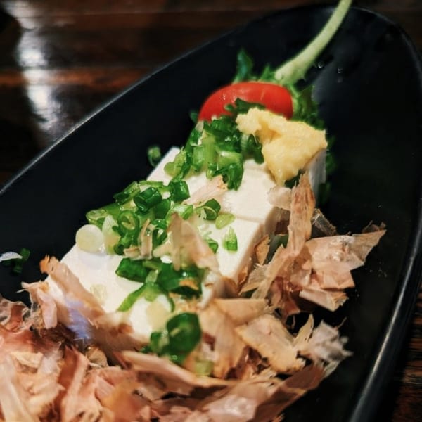 Japanese cold tofu topped with green onions, ginger, tomato, and bonito flakes in a black dish.