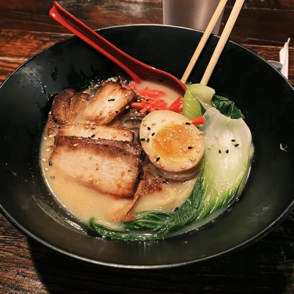 A black bowl of Ginya Izakaya ramen with pork, egg, bok choy, and red ginger on a wooden table.